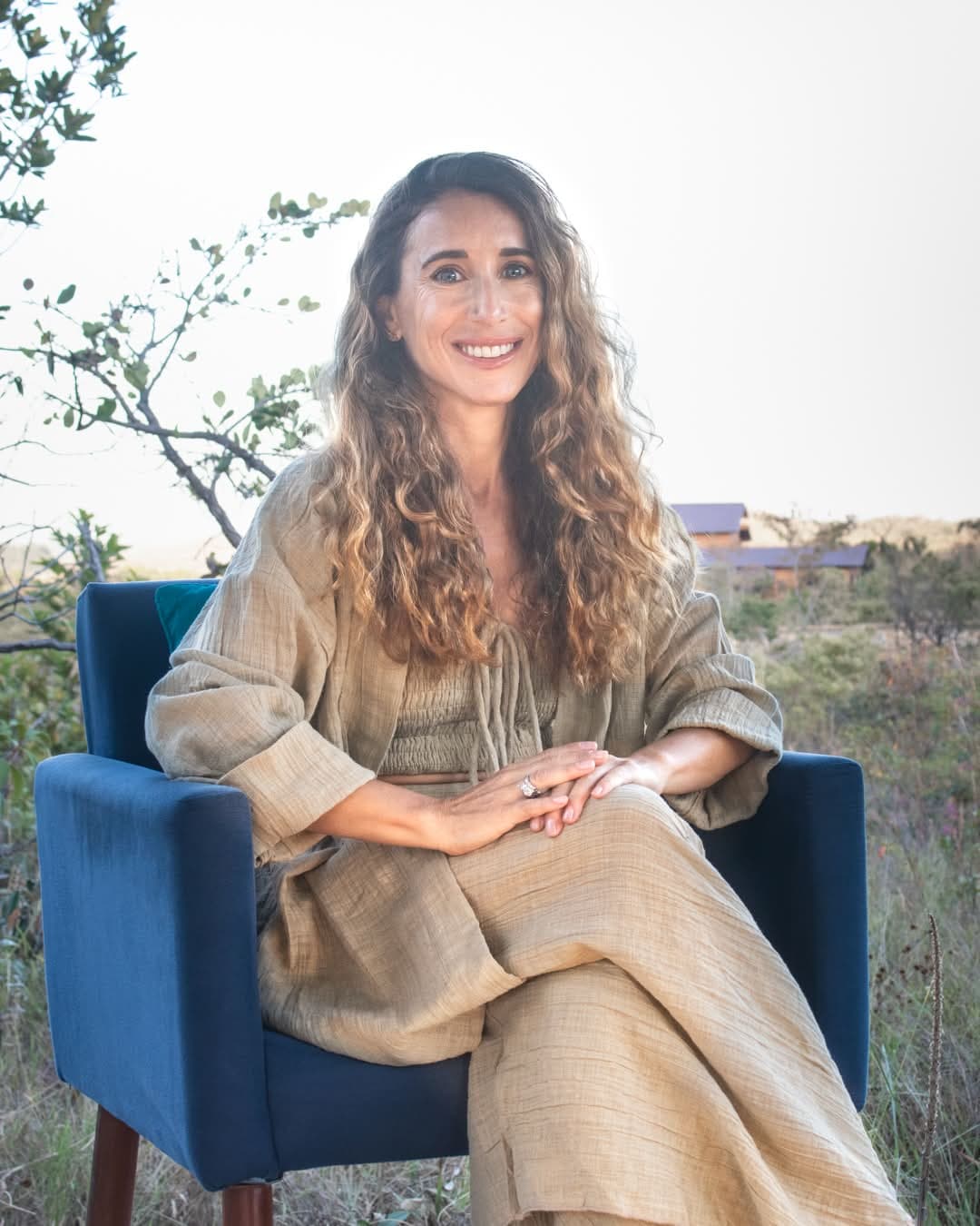 Jennifer Brooke Lawless, smiling, seated in a blue chair outdoors in linen at golden hour